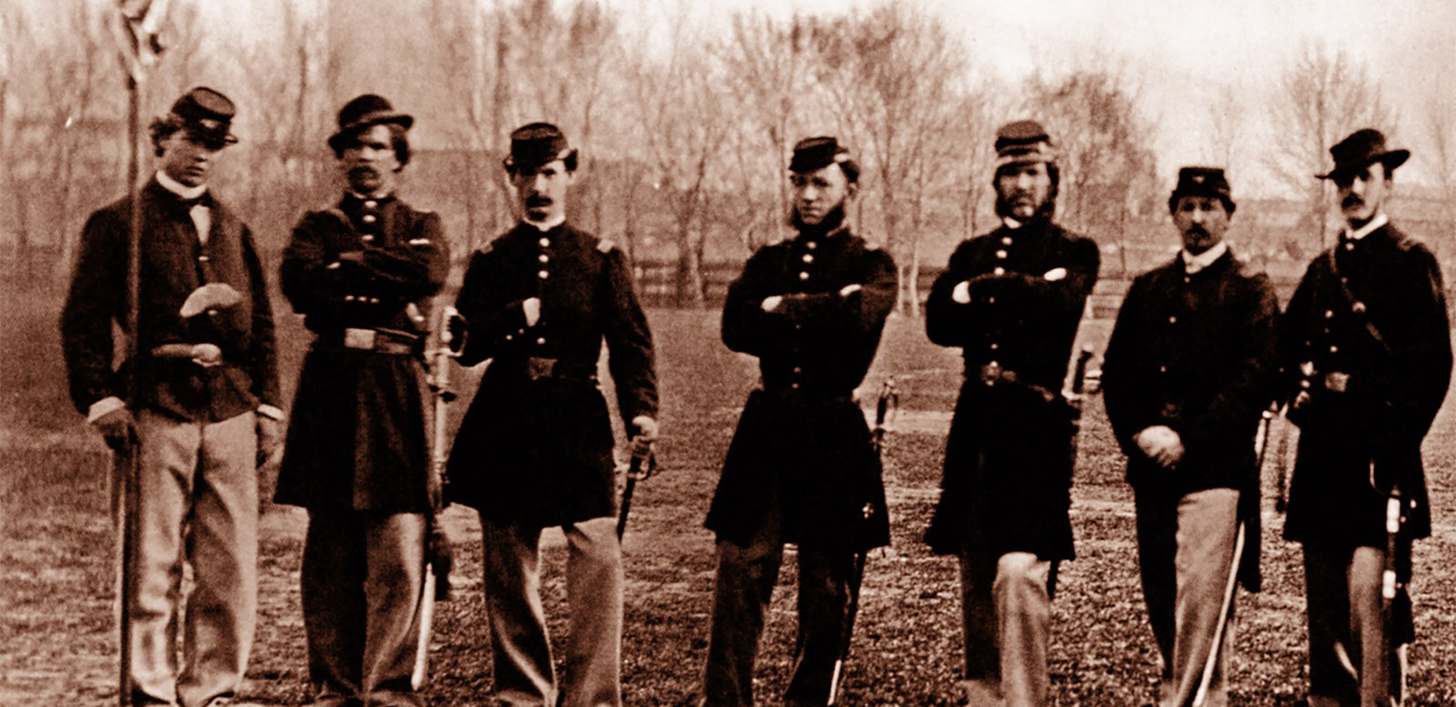 Black and white photograph of a row of men in union army uniforms standing in front of the unfinished Washington Monument.
