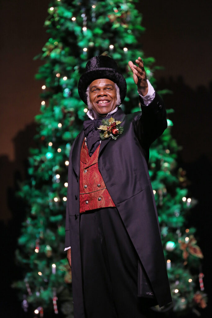 A smiling man in a black velvet top hat, red satin vest, tuxedo jacket with a festive holly and evergreen boutonniere, stands in front of a Christmas tree.