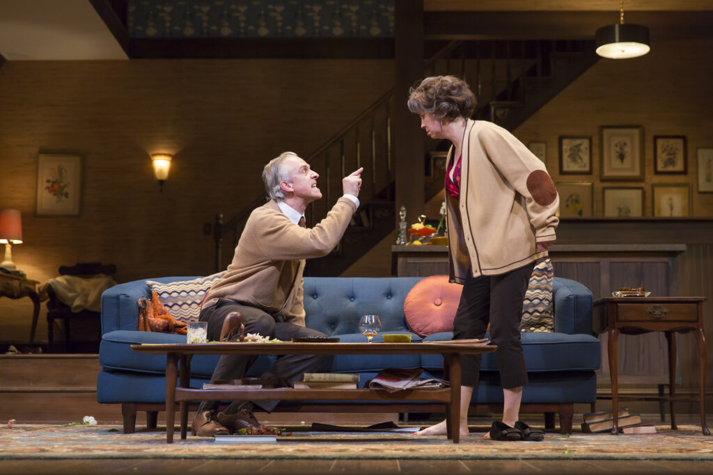 An older man sits on a couch and speaks angrily to an older woman who stands over him.