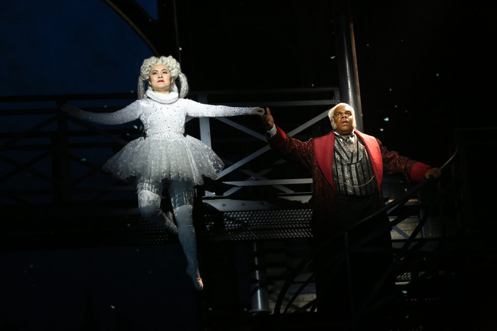 A spirit flies through the air in a sparkling, long-sleeved unitard and matching tutu. She grasps the hand of Ebenezer Scrooge and the two glide upward.