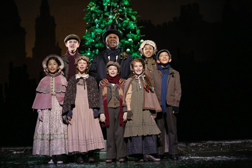 A smiling man in Victorian-style clothes and a top hat stands with a group of children, also in Victorian-style clothing, in front a large Christmas tree.