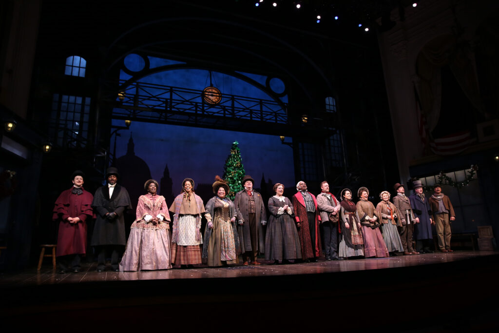 Fifteen people stand on stage in assorted Victorian costumes. The Presidential Box is seen above the stage, dressed with festive evergreen swags and lights.