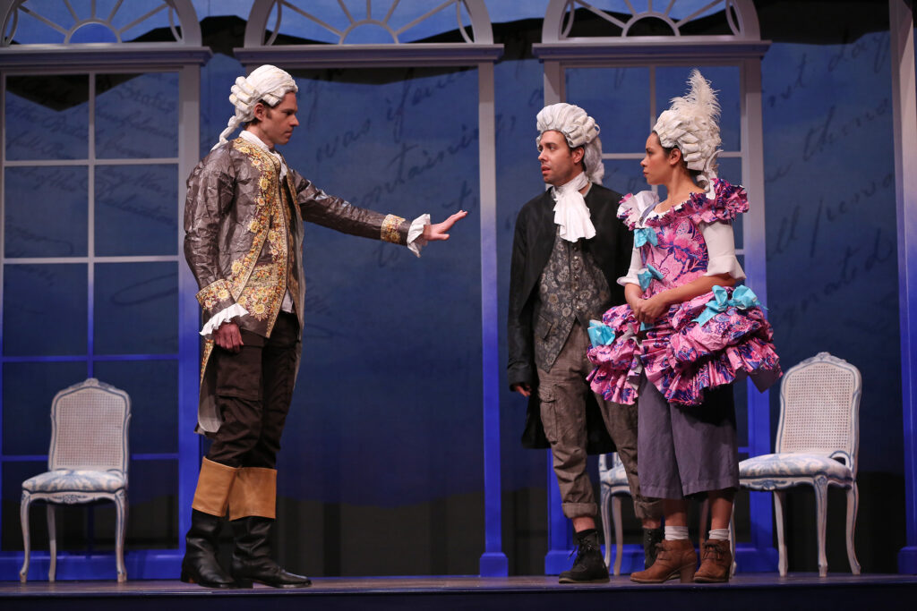 An older free man stands at left with his hand out, blocking the passage of an enslaved man and woman. The older man wears an 18th-century coat and vest in brown with gold flowers, a white wig and casual contemporary pants. The other man wears an 18th-century white wig, a black coat, a gray vest with flowers and casual contemporary pants. The woman wears an 18th-century white wig, a bright pink dress with a blue floral design and casual contemporary pants. Both the clothes and wigs are made of paper. They stand in front of a larger-than-life projection of the words from the Declaration of Independence.