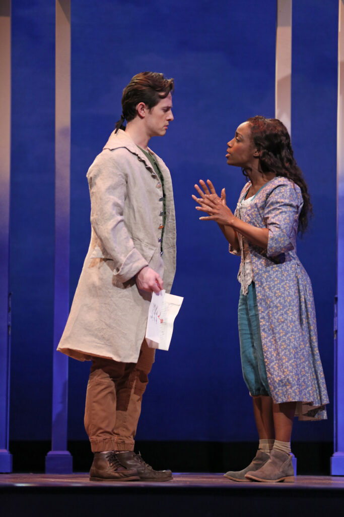 A man dressed in a plain tan linen coat looks mournfully at his former lover, a woman who was previously enslaved. He holds a letter. She wears an 18th-century-style purple dress with yellow flowers over casual contemporary blue capris.