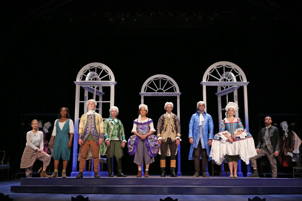 Nine actors stand on a raised platform in a line. Six of them are dressed in ostentatious 18th-century clothing and wigs made of paper. The other three wear more casual clothing inspired by the 18th-century. Behind them are white arched frames, representing mansion windows and doors.