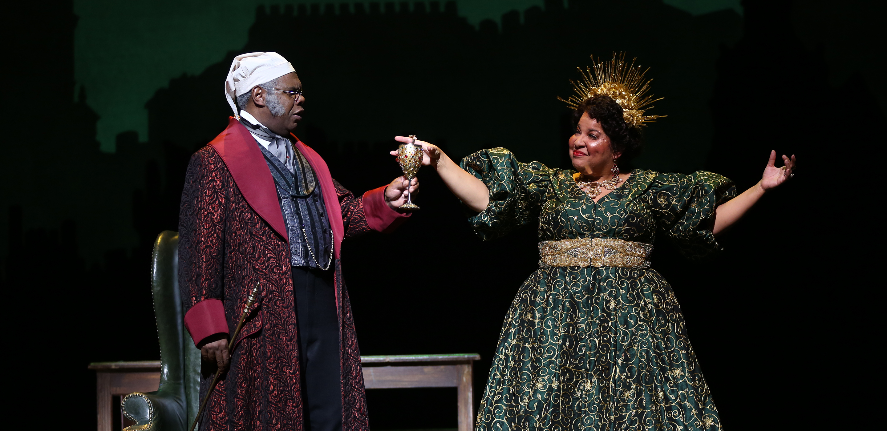 A man dressed in a Victorian robe and nightcap holds a gold goblet in a toast. He stands next to woman dressed as Spirit of Christmas Present in a richly decorated green Victorian-style dress and sunburst crown.