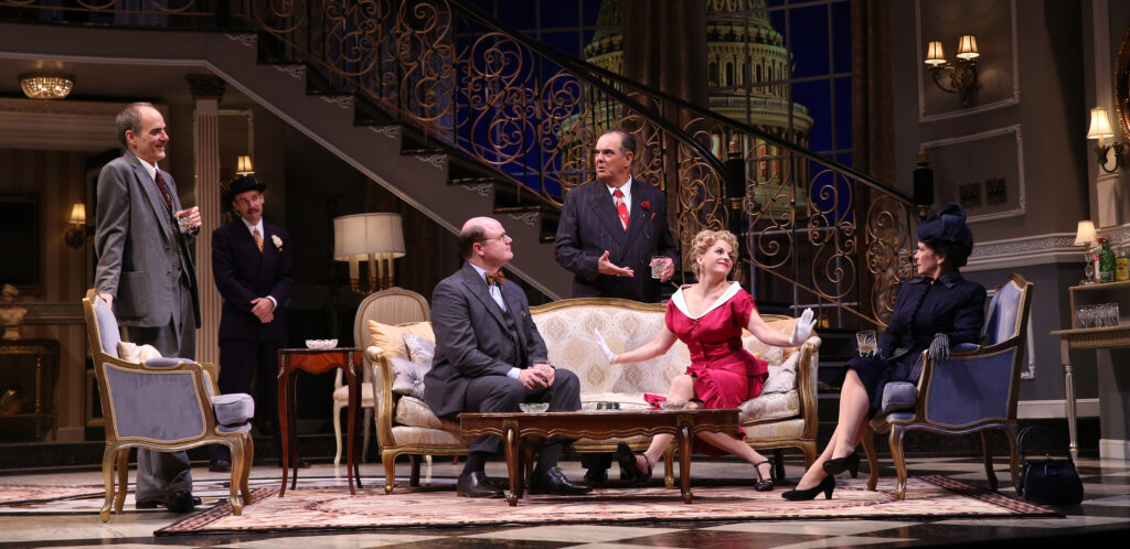 On a set designed to look like a fancy hotel living room in the 1950s, a woman in a red dress and a man in a three-piece suit sit on a sofa. Four others stand around them.