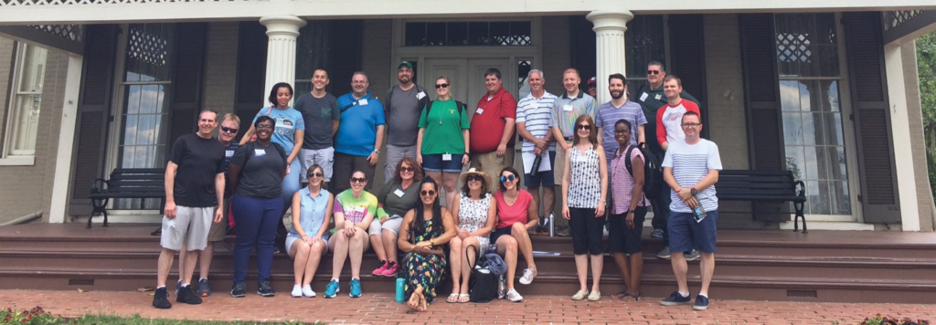 24 teachers arranged in two rows smile and pose for a group photo on the steps of Frederick Douglass National Historic site. Behind them is a large, Victorian house with a wide front porch with lattice trim, and large, floor to ceiling windows on either side of the front door.