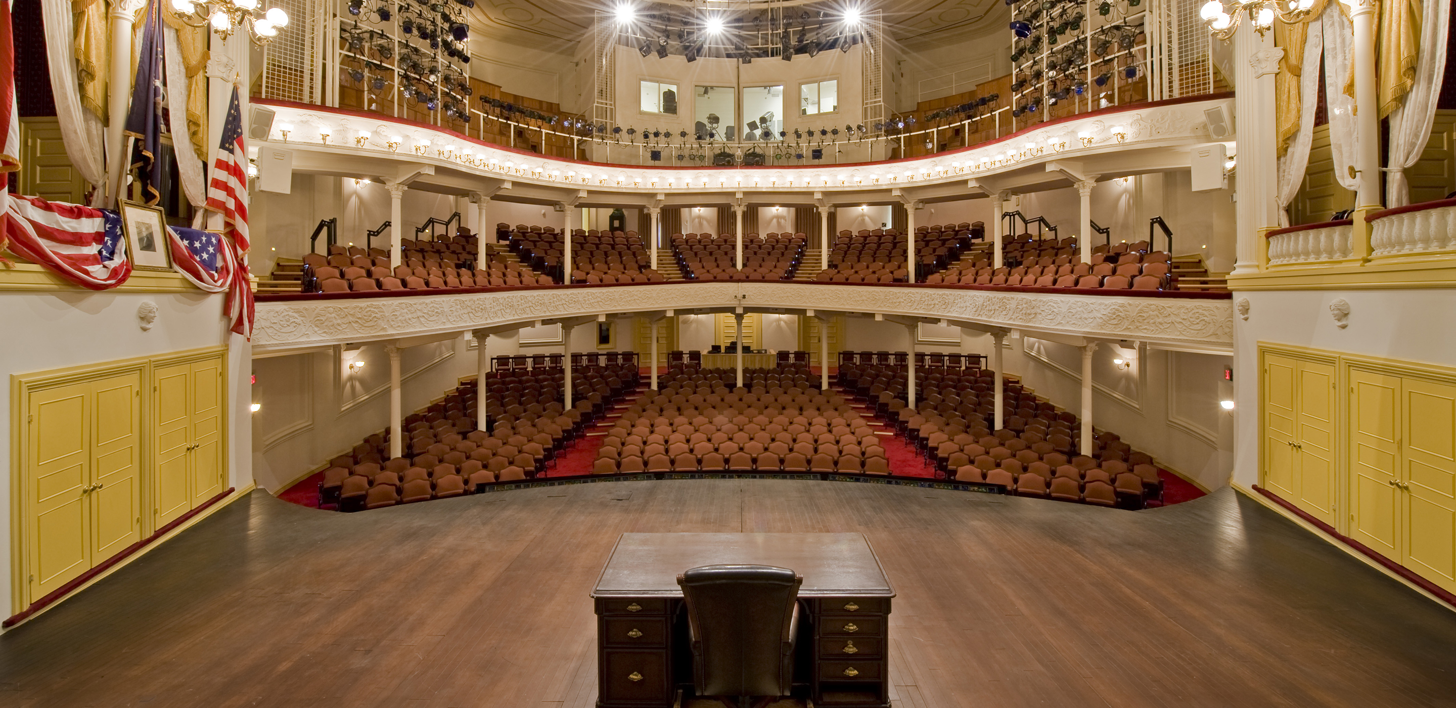 The view from the Ford’s Theatre stage looking out to the audience. To the left of the stage is the President Box with an American flag, a framed picture of George Washington and American flag bunting draped over the box. To the right is another box with yellow and white curtains. In the center of the stage is a wooden desk. The view includes two levels of seating and rows of lighting equipment on the third level.