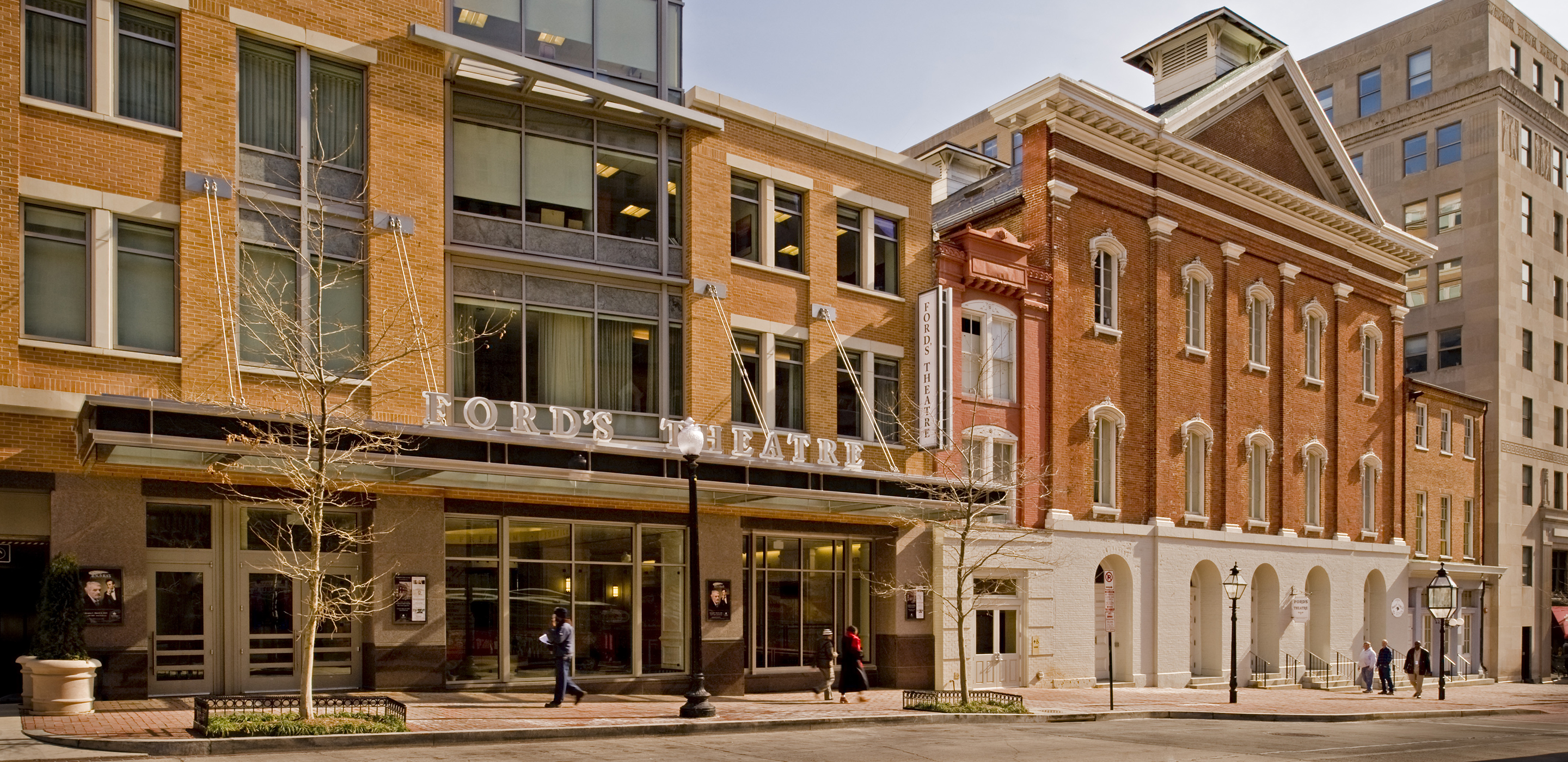 A street view of the lobby and the historic Ford’s Theatre. The lobby, on the left, is a modern building with large glass windows and a large sign reading “Ford’s Theatre.” The historic theatre, to the right, is a three-story brick building with a series of arched entrances.