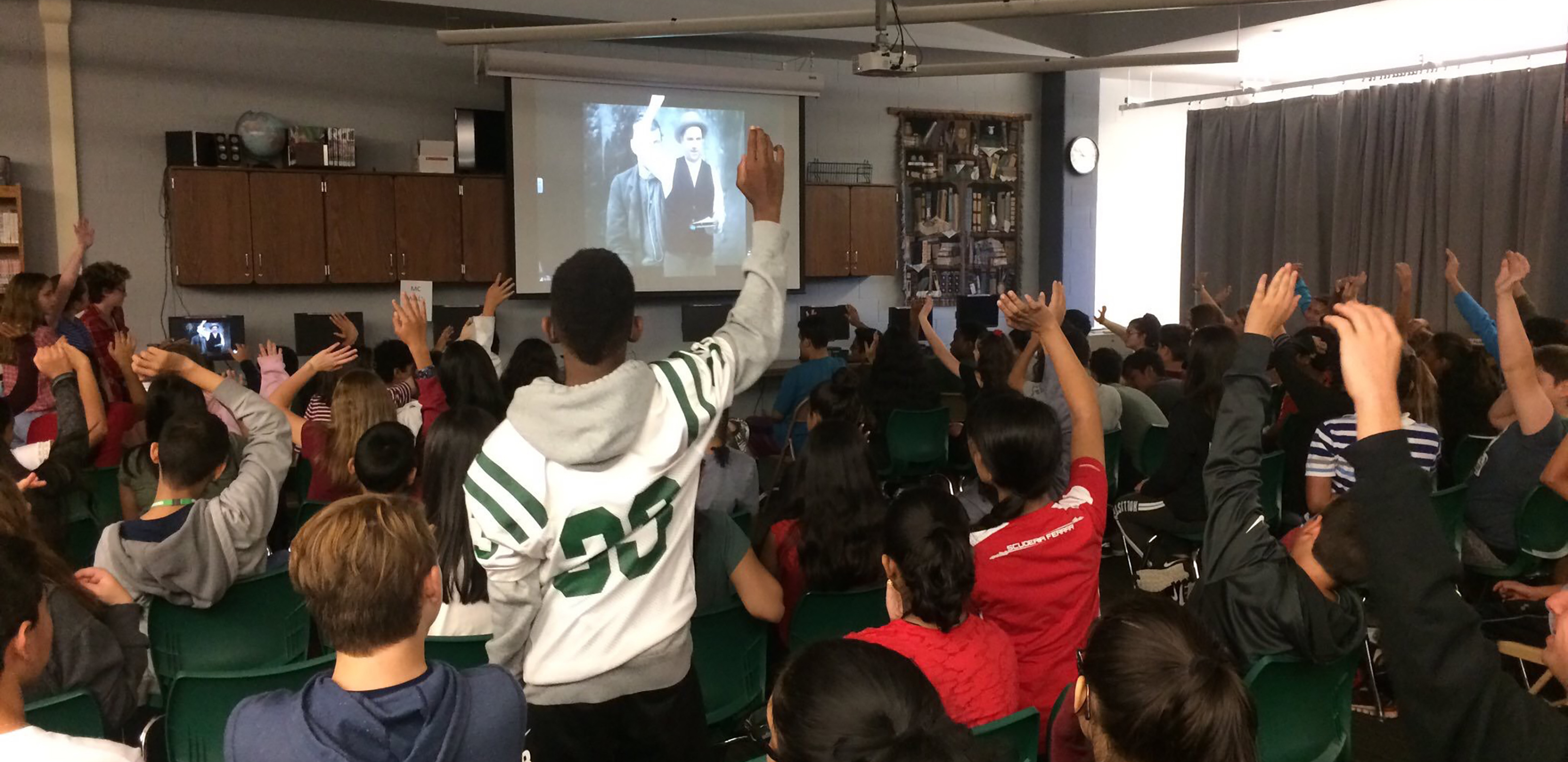 A large classroom full of students watching an actor portraying Detective McDevitt on a projector screen. Many students are raising their hands to participate.