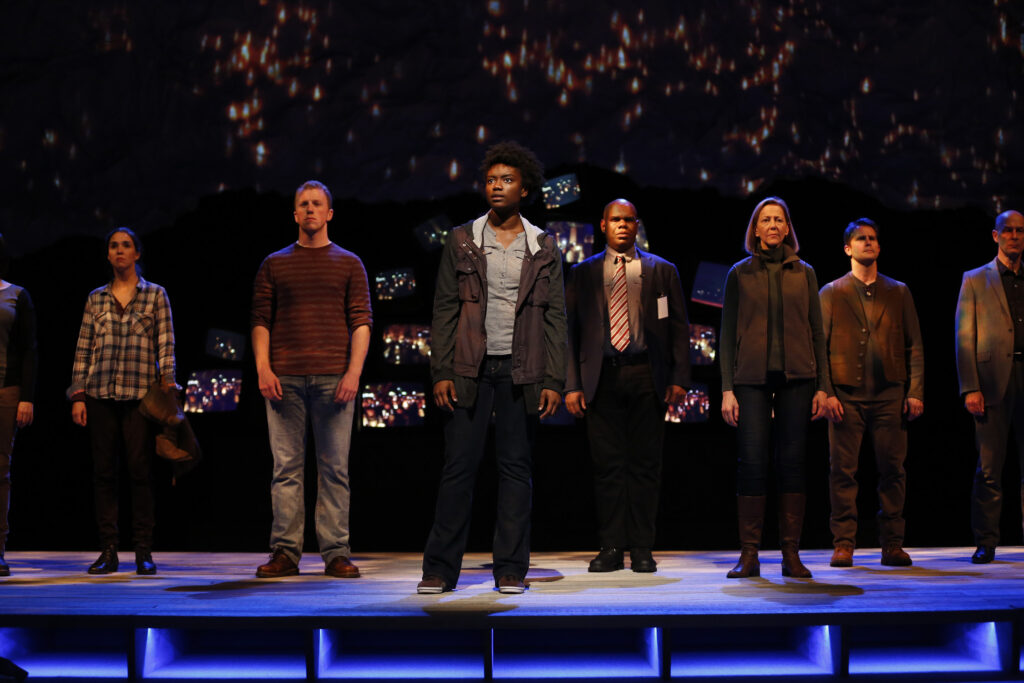 Eight people stand on a stage and look out at the audience. Behind them a jumbled pile of television screens all show images from a candlelight vigil.