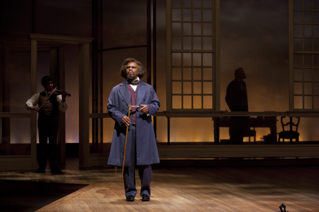 An actor portraying Frederick Douglass holds a cane and stands on a stage. Behind him a violinist plays.