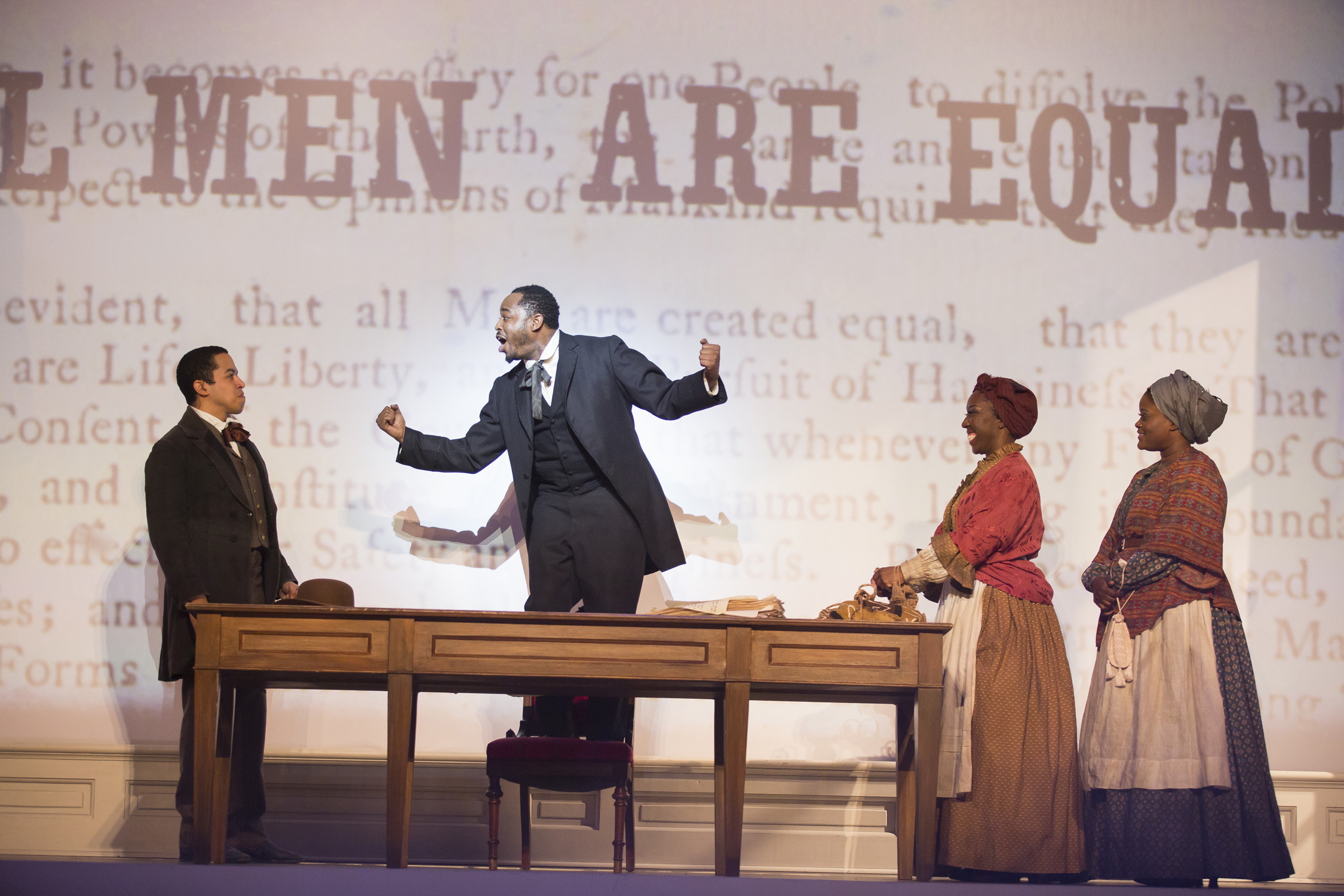An African-American man stands on a chair behind a table and gives a speech. Two women and a man listen to him. Behind him the text of the Declaration of Independence is projected, with the words "All Men Are Equal" superimposed.