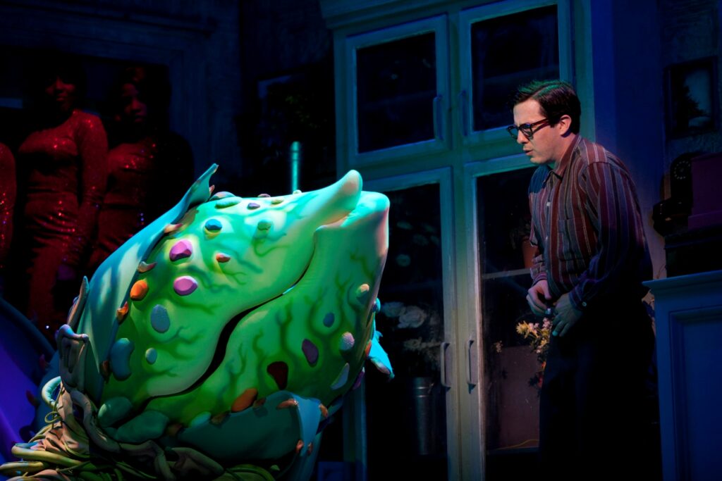 A man in glasses and a button down shirt stands in a bedroom and talks to a giant plant with a huge mouth.
