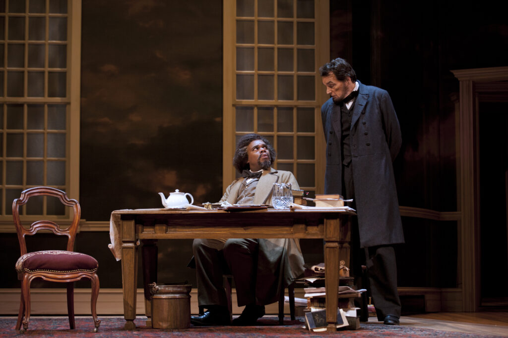 An actor portraying Frederick Douglas sits at a cluttered desk while an actor portraying Abraham Lincoln stands over him and speaks.