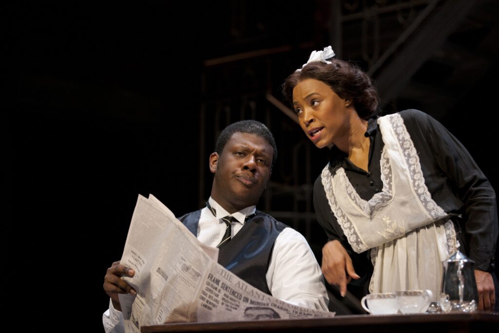 An African-American man in a suit reads an newspaper and speaks to a woman in a maid's outfit.