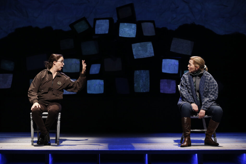 Two women sit in chairs some distance apart on an empty stage. One, who wears a police uniform, raises a hand and speaks to the other. In the background is a jumbled pile of staticky television screens.