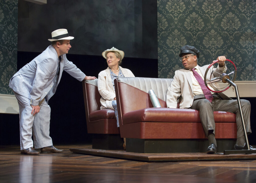 An actor in a light suit and panama hat leans over to address two actors in a “car.” An actress in a hat, cardigan and pearls sits in the back bench seat. An actor in a suit and chauffeur’s cap sits in the front bench seat with his hand on a steering wheel.
