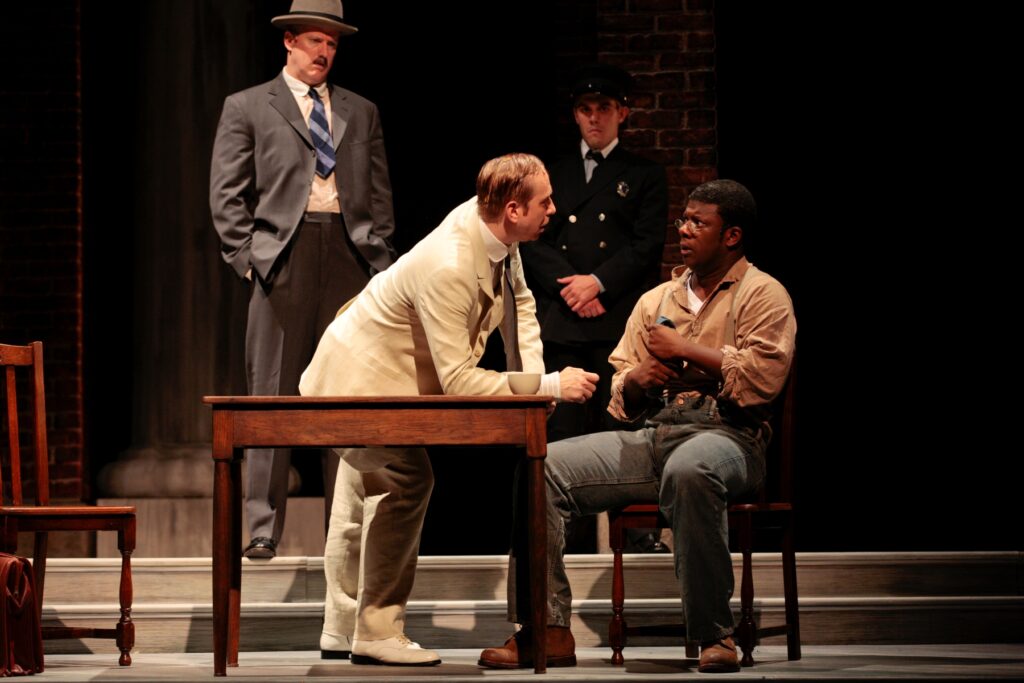 An African-American man in work cloths sits in a chain and looks nervously at a white man in a white suit who leans over a table and speaks to him. A policeman and a man in a suit stand behind them.