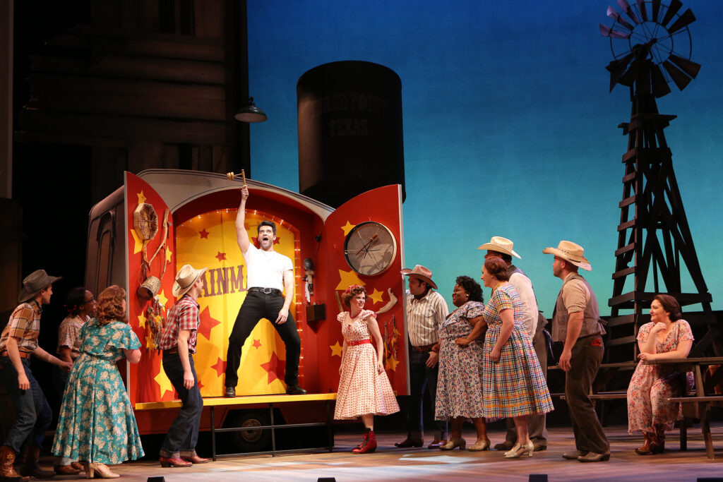 Men and women gather around a brightly painted automobile trailer. The back doors of the trailer are open, and inside a man sings and dances.