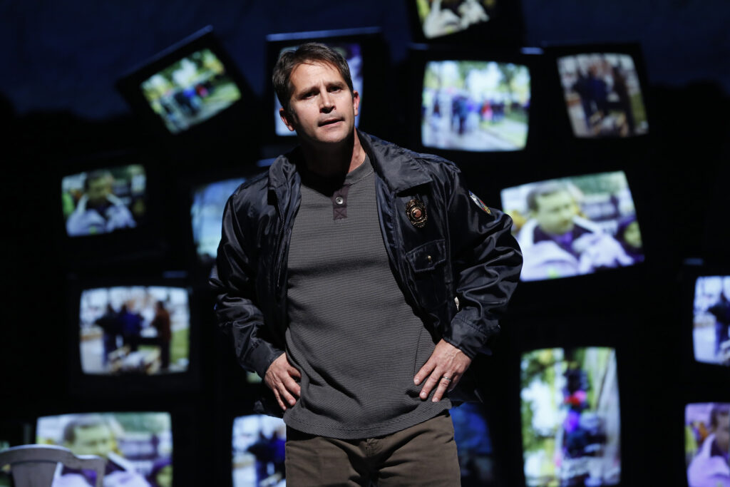 A man wearing a police jacket stands in front of a jumbled pile of television screens, all displaying blurred pictures.