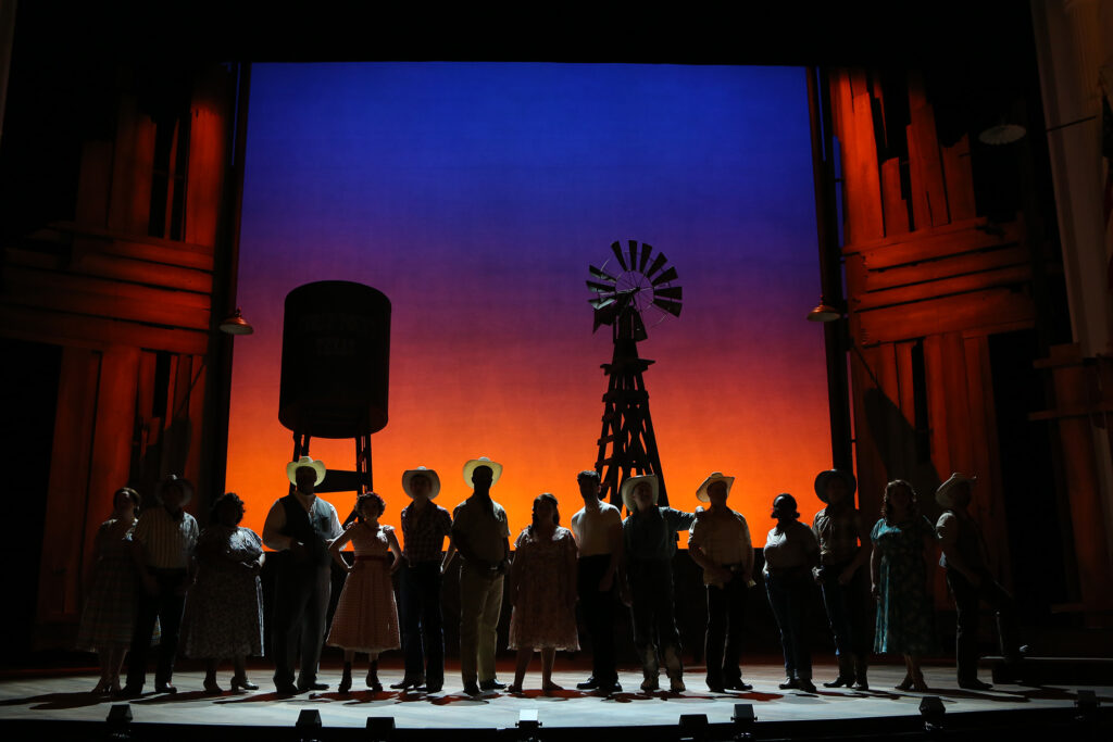 A group of men and women stand in shadow and look at the camera. Behind them the stage light resembles a sunrise.