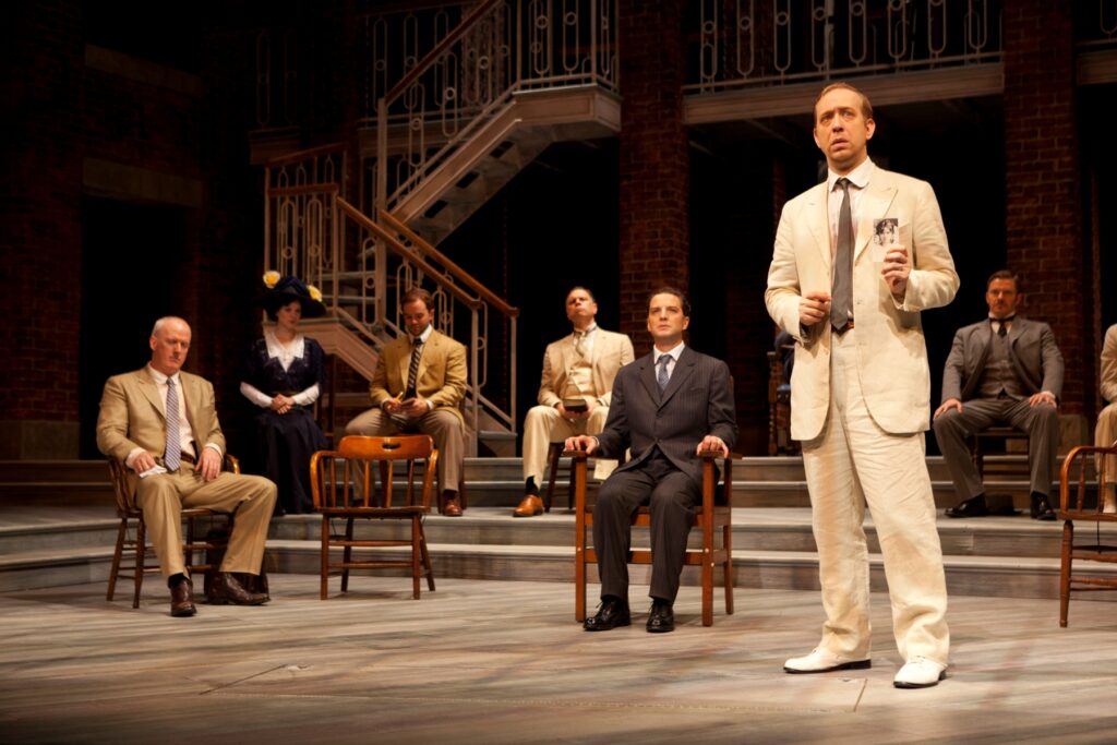 A group of men and women sit in chairs on a stage while a man in a white suit stands in front of them and holds up a photograph of a woman.