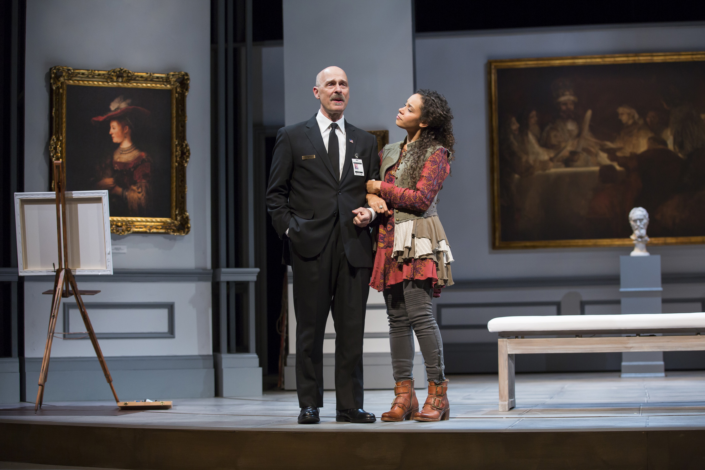 A man dressed as a security guard stands in a room in an art museum. A young woman is arm-in-arm with him and listens to him speak.