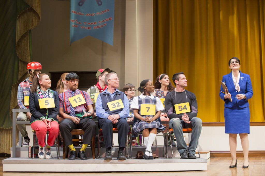 A group of 10 actors dressed as children sit in chairs on a stage, each with a number on a placard around their necks. A woman in a blue blazer stands next to them and speaks.