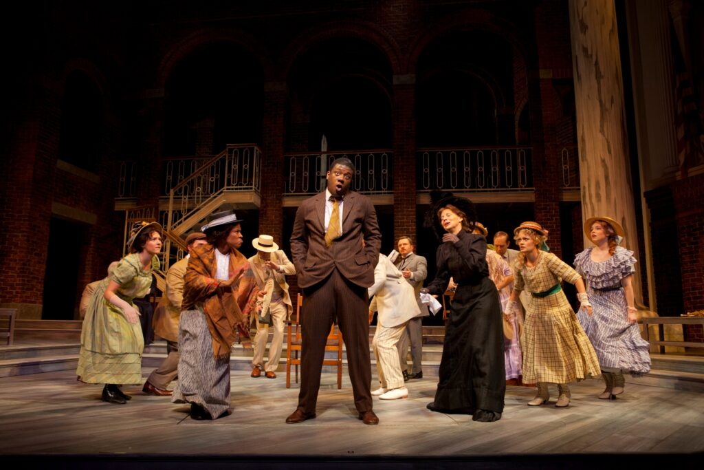 An African-American man in a suit stands on a stage and sings while a group of men and women dance around him.