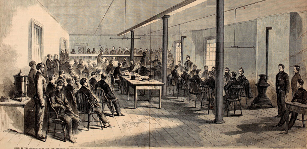 Drawing of a crowded courtroom. The defendants sit at a table in the center of the room, surrounded by guards and onlookers.