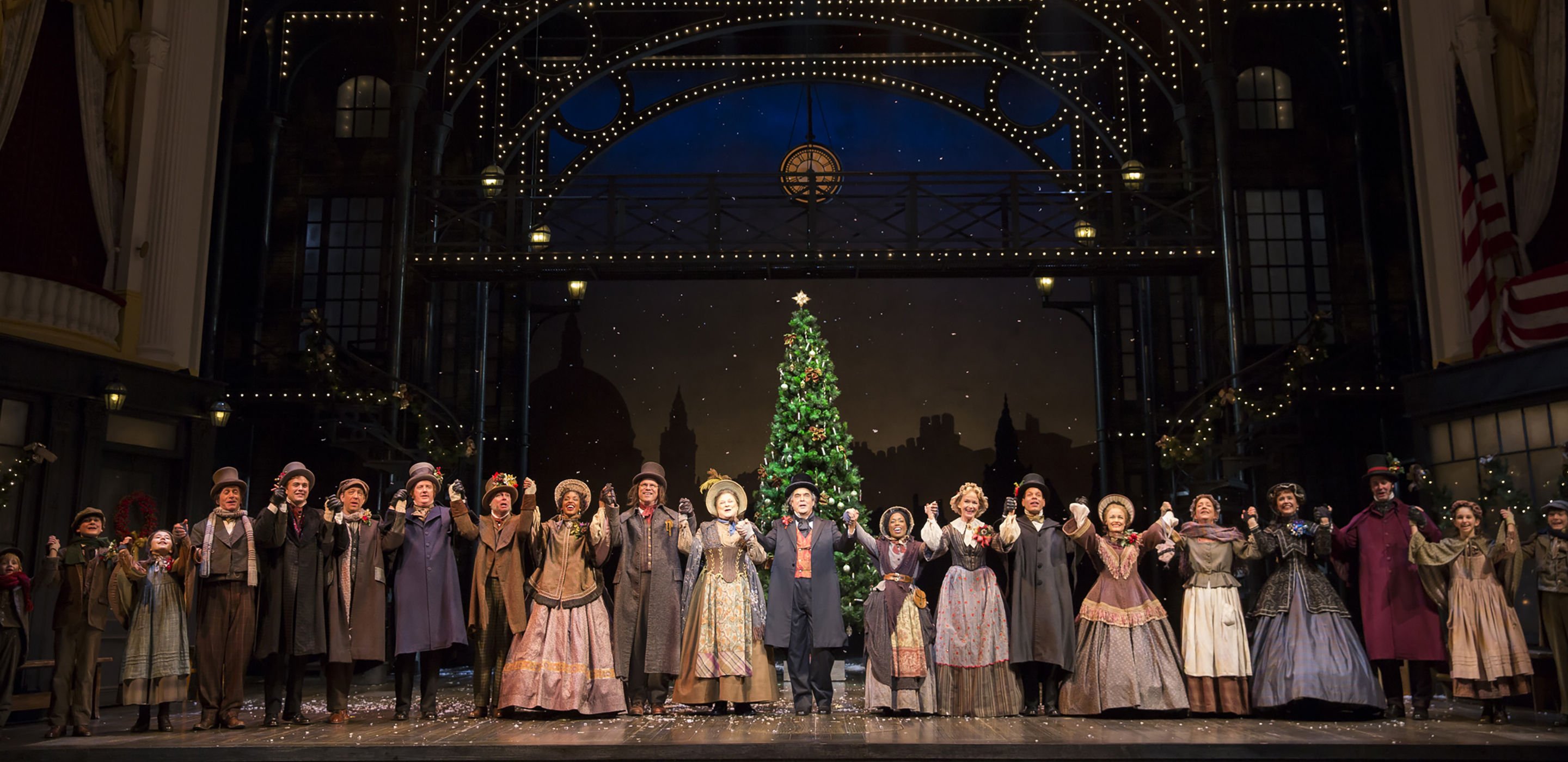 A large group of men and women in Victorian era clothing stand in a line hand-in-hand on a stage and face the audience. Behind them is a Christmas tree.