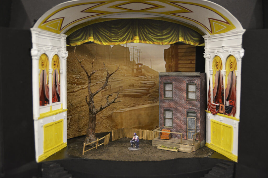 Miniature scaled model of the Ford’s Theatre proscenium stage with the set design for “Fences”. Set features a two-story brick façade of a row home, small white fence surrounding a dirt yard. In the neighbor’s yard is a tall, established but barren tree.