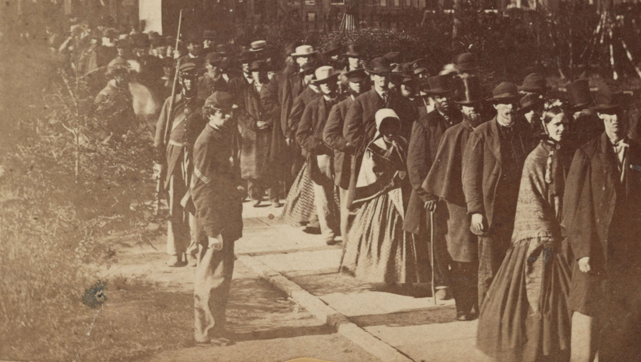 Sepia photograph of of a long line of people in mourning clothes. To the right is a city park, and behind them are a row of buildings.
