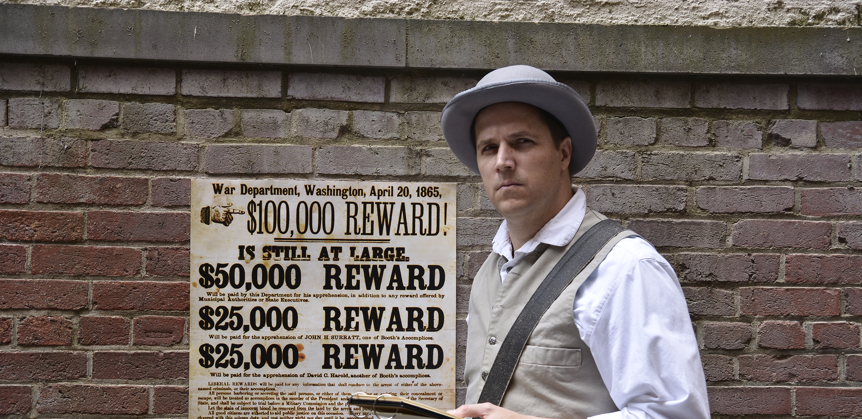 An actor stands dressed as an 1860s detective, holding a notepad. Behind him on a brick wall is posted a reward poster for the capture of Lincoln's assassin.