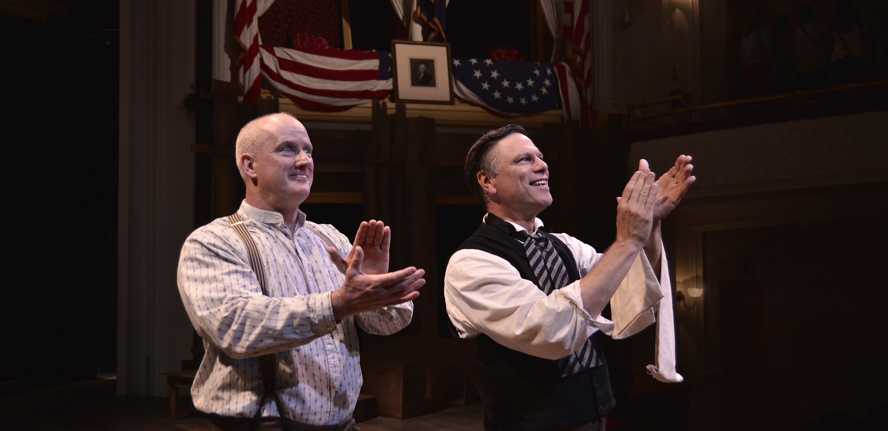 Actors playing Harry Hawk and Harry Ford wear 1860s costumes and mourning arm bands. They stand smiling and clap at someone not pictured in the distance. Behind them is the flag-draped Presidential Box at Ford’s Theatre.