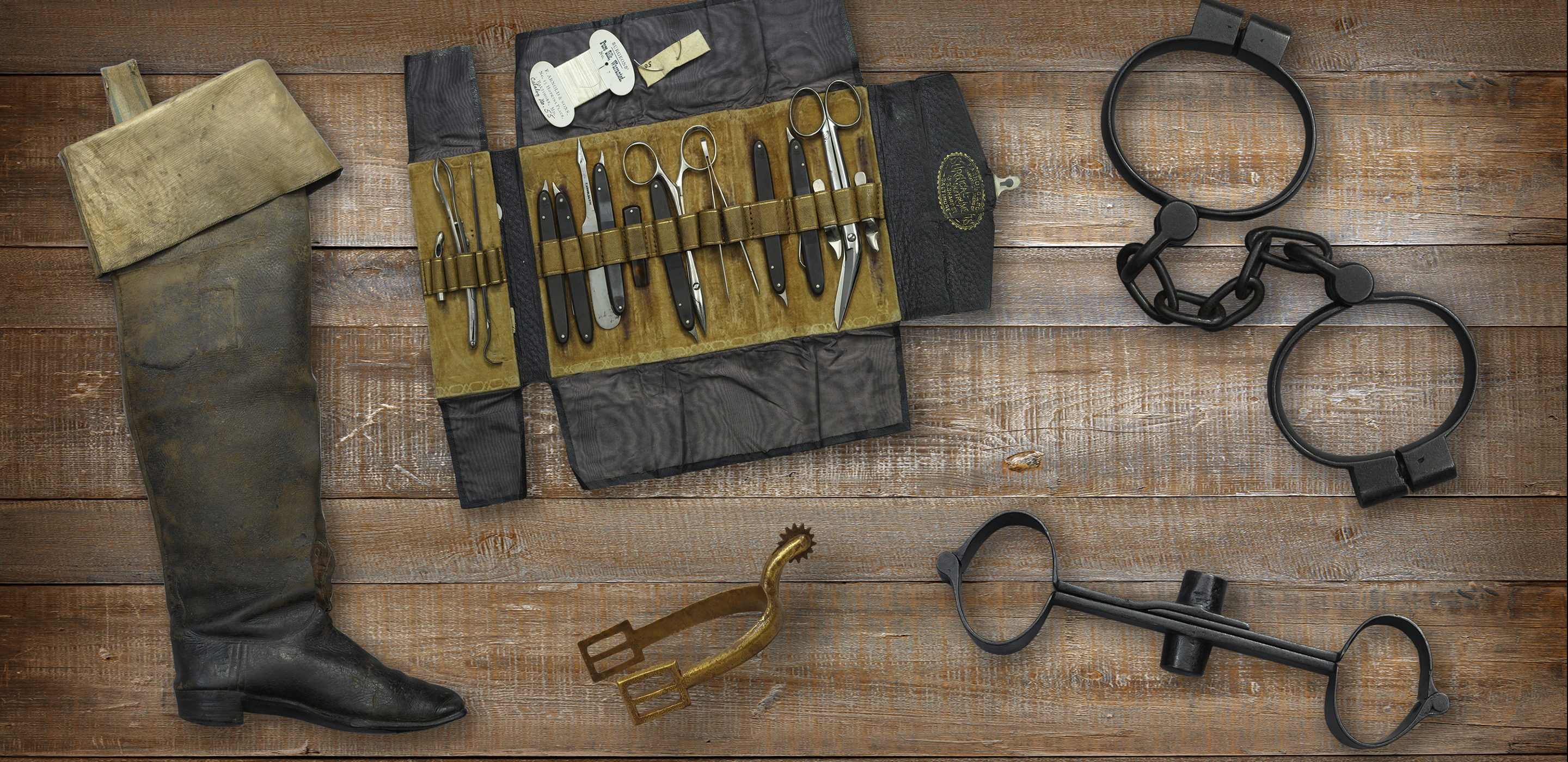 A photograph of a number of items on a wooden tabletop: A boot, a set of surgical instruments, a set of handcuffs, a set of ankle shackles and a spur.