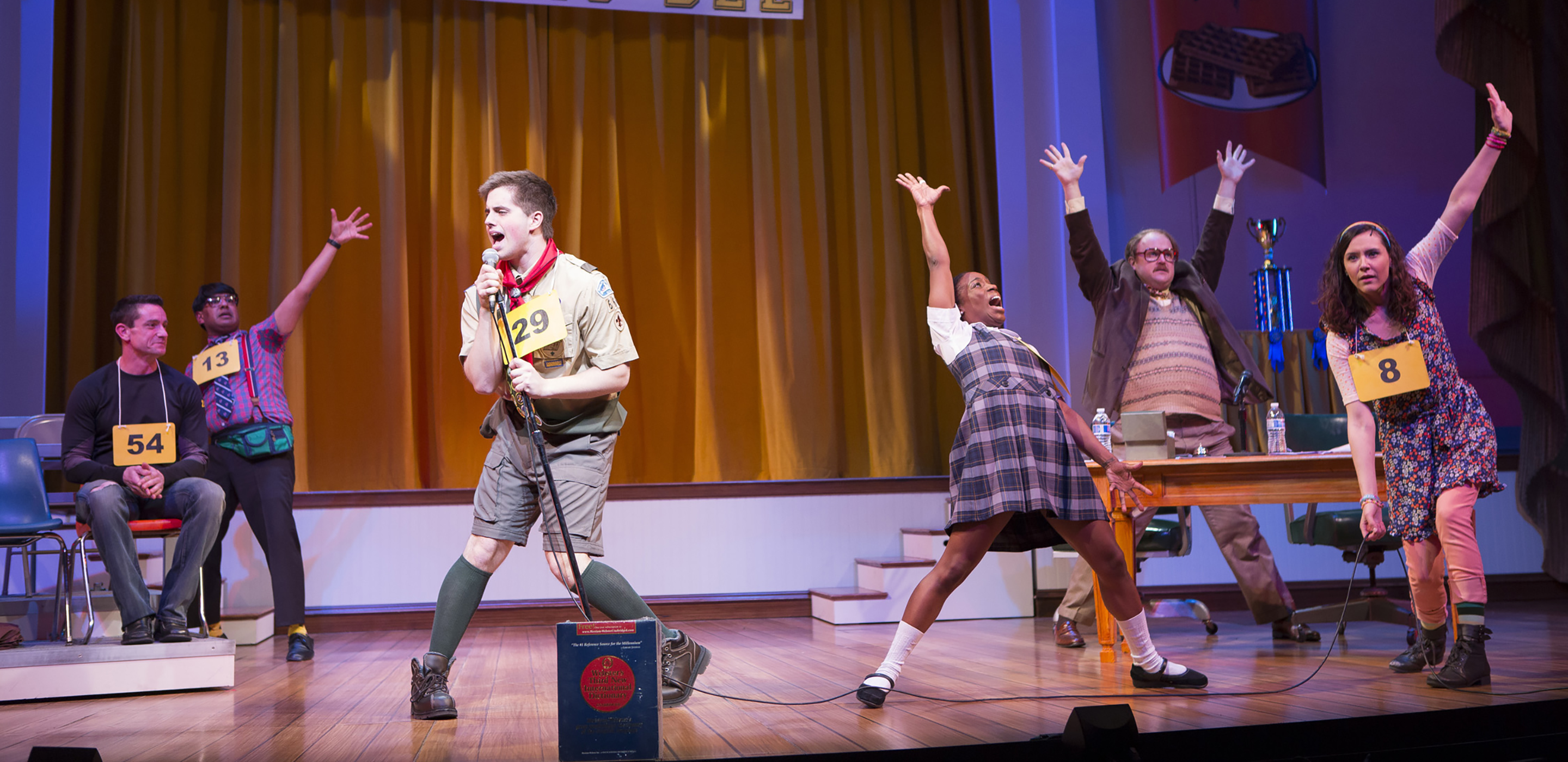 An actor in a boy scout uniform sings into a microphone on a stage. He has a placard around his neck with the number 29 written on it. Other students and teachers dance around him.