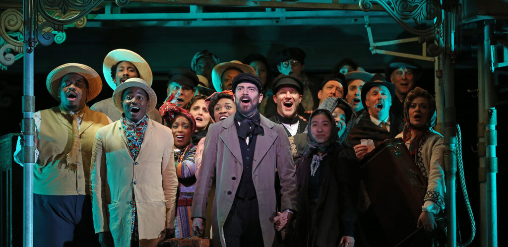 A large group of people in travelling outfits stand together under scaffolding and sing.