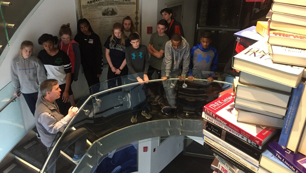 A group of students gather on a spiral staircase, while a Ford’s Education staff guide shows them the Lincoln Book tower, a giant column of books in the center of the staircase.