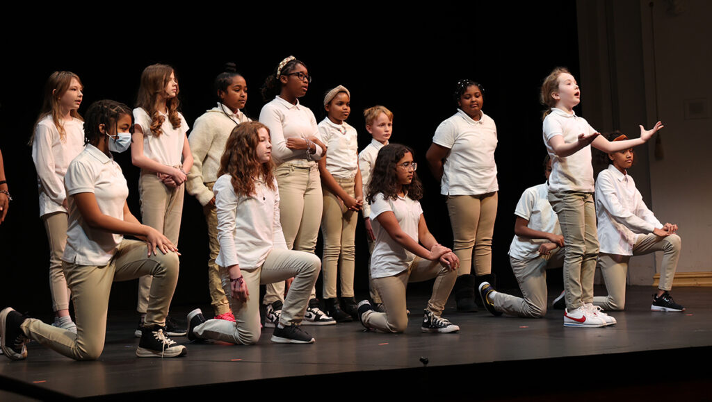 A group of schoolchildren in white shirts and tan pants stand or kneel on a stage.