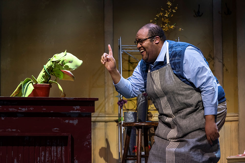 A Black man smiles and raises his finger in front of a small green strange and interesting plant.