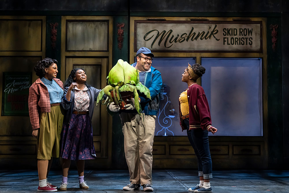 Three Black women in colorful streetwear surround a smiling Black man, who is holding a medium green strange and interesting plant in a pot. They stand in front of a sign that reads “Mushnik Skid Row Florists.”