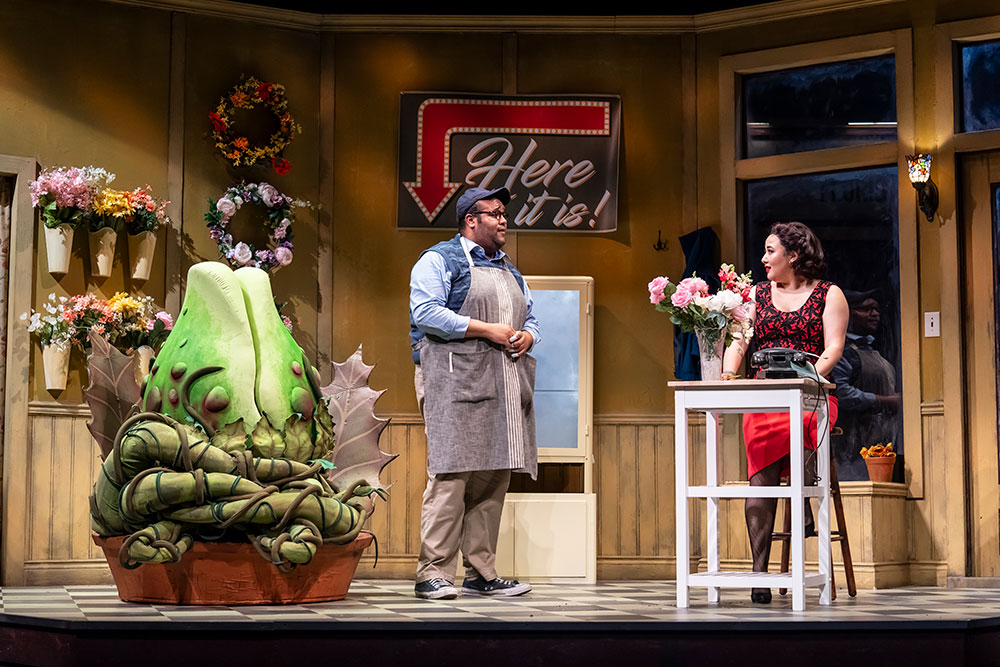 A Black man wearing a gardener’s apron and an Asian person wearing a red dress with black fabric chat in a plant shop, with a large green strange and interesting plant in a pot to their left. A sign pointing to the plant reads “Here it is!”