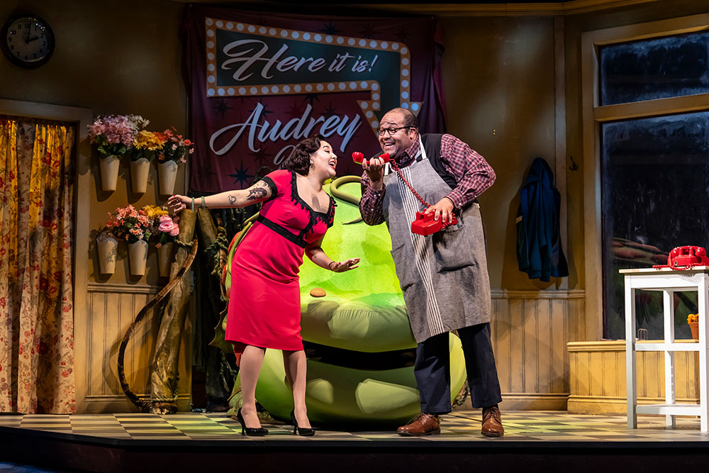 An Asian person wearing a red dress and a Black man wearing a gardener’s apron stand in front of a large, green strange and interesting plant. The Black man is holding a red 60’s era telephone.