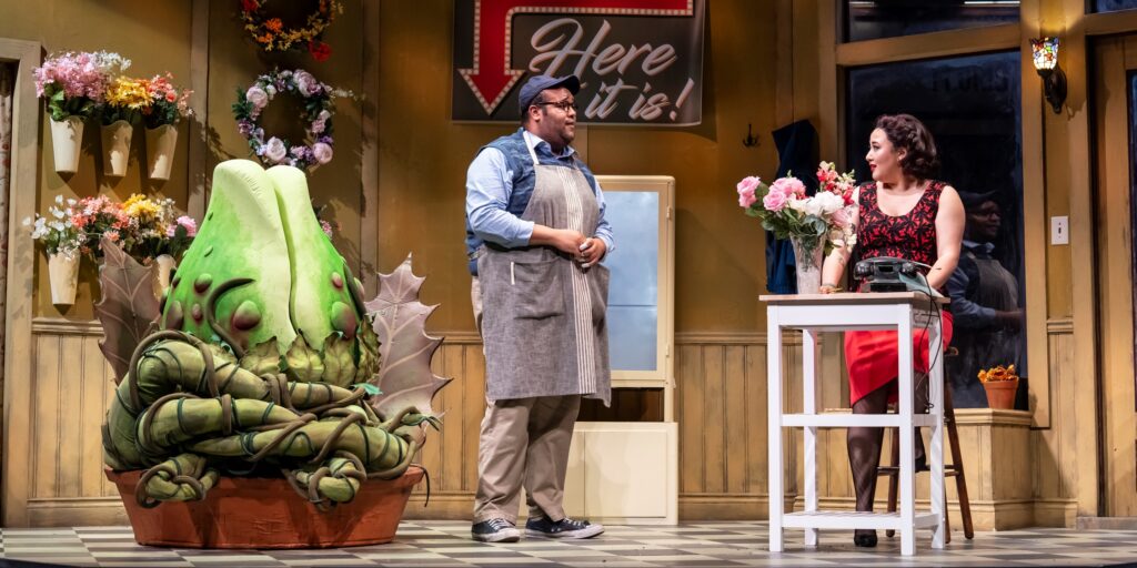 A Black man wearing a gardener’s apron and an Asian person wearing a red dress with black fabric chat in a plant shop, with a large green strange and interesting plant in a pot to their left. A sign pointing to the plant reads “Here it is!”