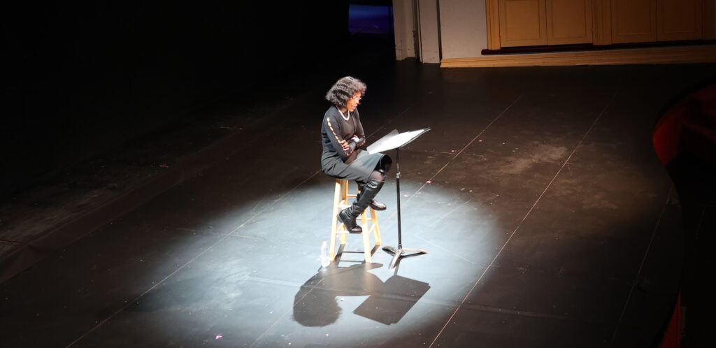 A Black woman in black clothing sits on a stage and reads from a script.