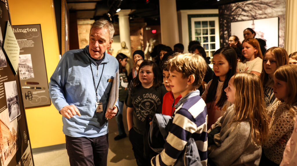 A group of children in a museum listen to an adult explain an exhibit.